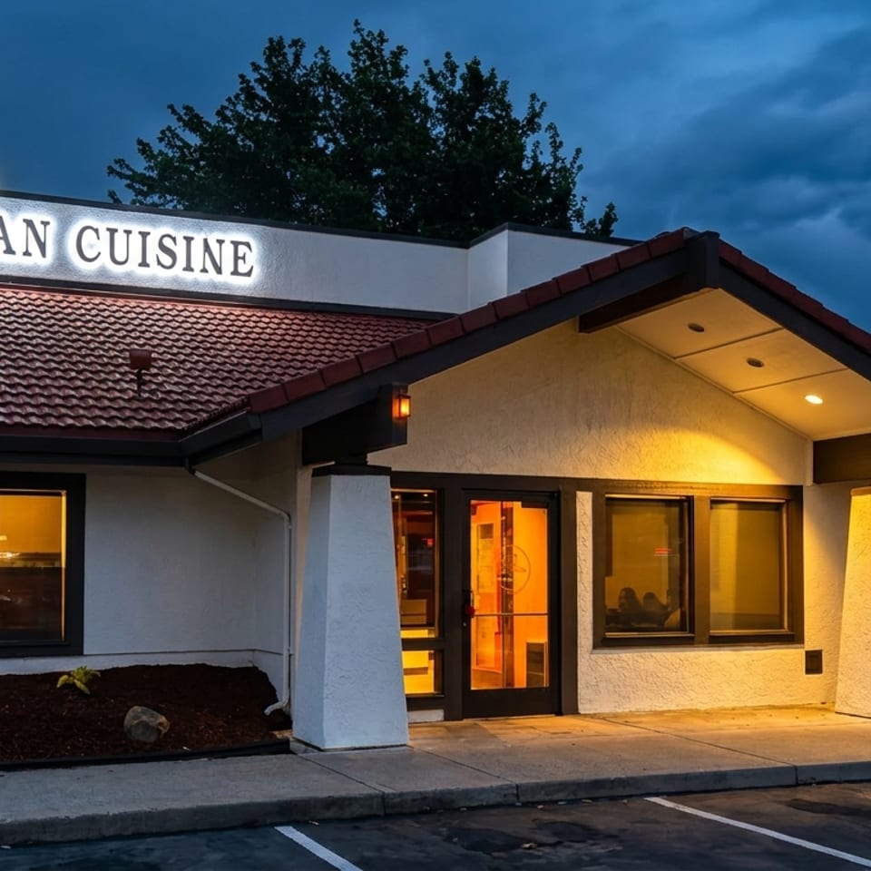 Indian cuisine restaurant exterior illuminated at dusk, with tiled roof, glowing windows, entrance canopy, and parking spaces.
